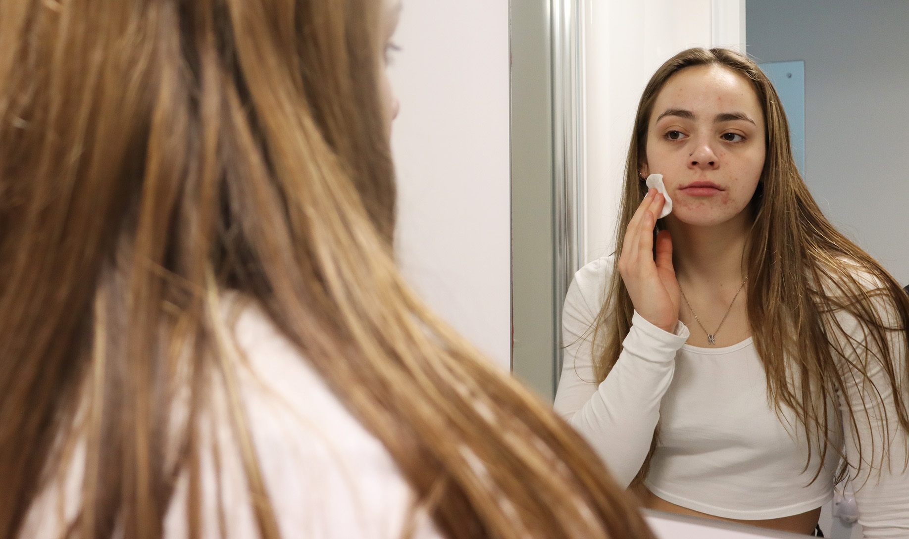 An image of a female looking into the mirror with a cotton pad on her face.
